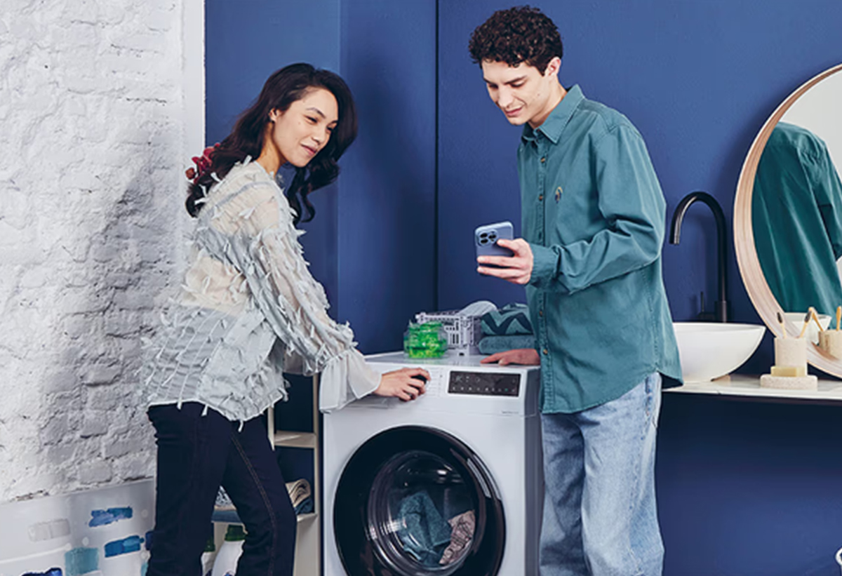 Απεικονίζεται το πλυντήριο σε ένα μπάνιο και ένας άντρας να εξηγεί σε μια γυναίκα την εφαρμογή hOn.